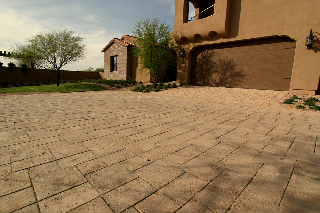 Tan rectangular concrete pavers laid in a running-bond pattern across a residential driveway in front of a house with a timber garage door
